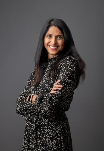 Rupa Bannerjee, a slender woman with long dark hair wearing a black and white specked dress against a rich charcoal grey background. She has her arms crossed and is smiling.